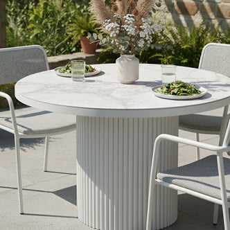 Evone Aluminium Outdoor Dining Table in White with round tabletop, fluted base, four white-metal frame chairs, vase with dried foliage, and plates of salad on a sunlit patio.