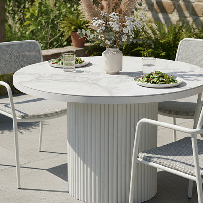 Evone Aluminium Outdoor Dining Table in White with round tabletop, fluted base, four white-metal frame chairs, vase with dried foliage, and plates of salad on a sunlit patio.