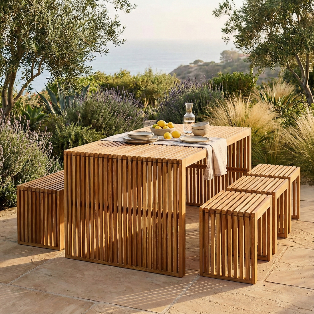 Grenfela 5 Seater Solid Timber Outdoor Dining Set featuring a slatted-wood table with matching stools on a stone patio, adorned with a linen runner, ceramic plates, lemons, and a water carafe.