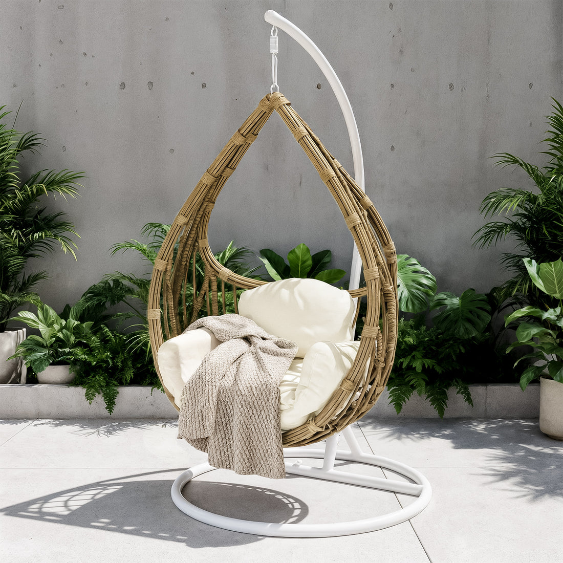 Hookipa Outdoor Rattan Egg Chair with Cushion in Cream White, suspended from a white stand, features an off-white cushion and knit throw, set on a patio with lush potted plants behind.