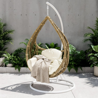 Hookipa Outdoor Rattan Egg Chair with Cushion in Cream White, suspended from a white stand, features an off-white cushion and knit throw, set on a patio with lush potted plants behind.