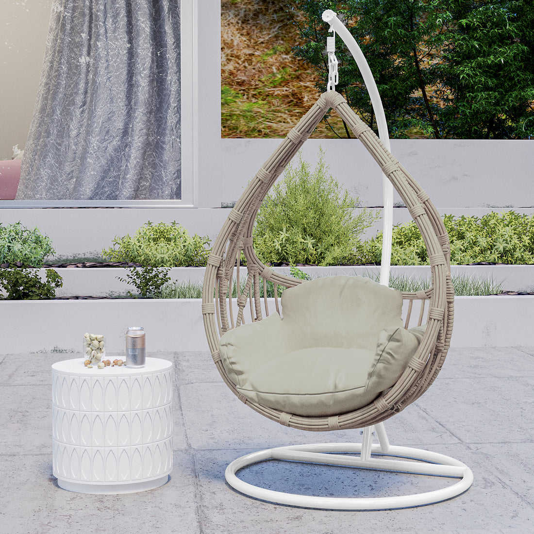 Hookipa Outdoor Rattan Egg Chair with Cushion in Sage, featuring a teardrop-shaped woven design suspended from a white stand, paired with a neutral cushion, next to a decorative white side table.
