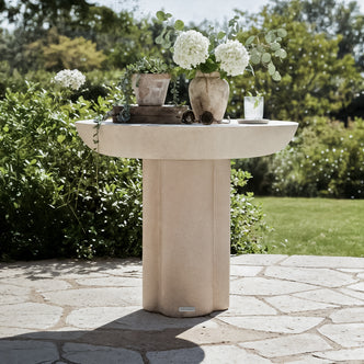 Jareth Round Concrete Outdoor Entry Table in Ivory on a patio, displaying a clay pot with succulents, a vase with hydrangeas and eucalyptus, and a glass with ice and mint.