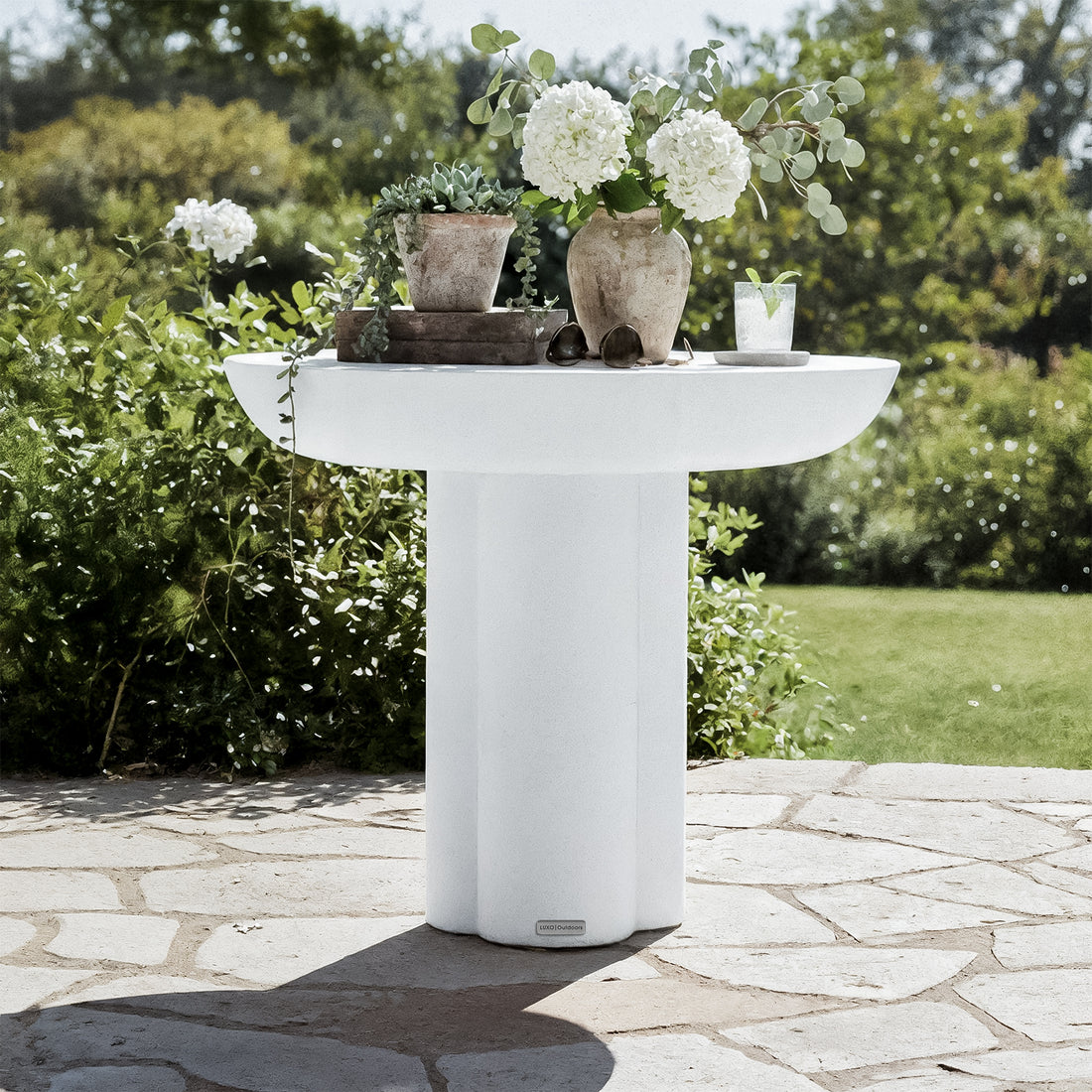 Jareth Round Concrete Outdoor Entry Table in White on a stone patio, adorned with terra-cotta vessels, wooden trays, sunglasses, and a drink, set against lush garden foliage.