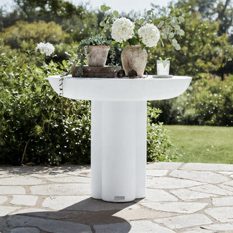 Jareth Round Concrete Outdoor Entry Table in White on a stone patio, adorned with terra-cotta vessels, wooden trays, sunglasses, and a drink, set against lush garden foliage.