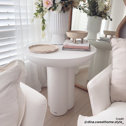 Jareth Round Concrete Outdoor Entry Table in White, featuring a sculptural base, topped with a scalloped tray, footed dish, hardcover books, and a ribbed vase with florals, amidst neutral-toned decor.
