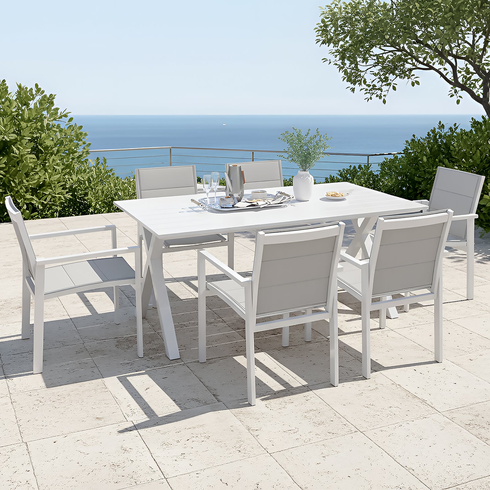 Jensen 6 Seater Aluminium Outdoor Dining Set in Light Grey features a sleek white table with matching armchairs, adorned with a ceramic vase, metal tray, and snacks, overlooking a serene ocean view.