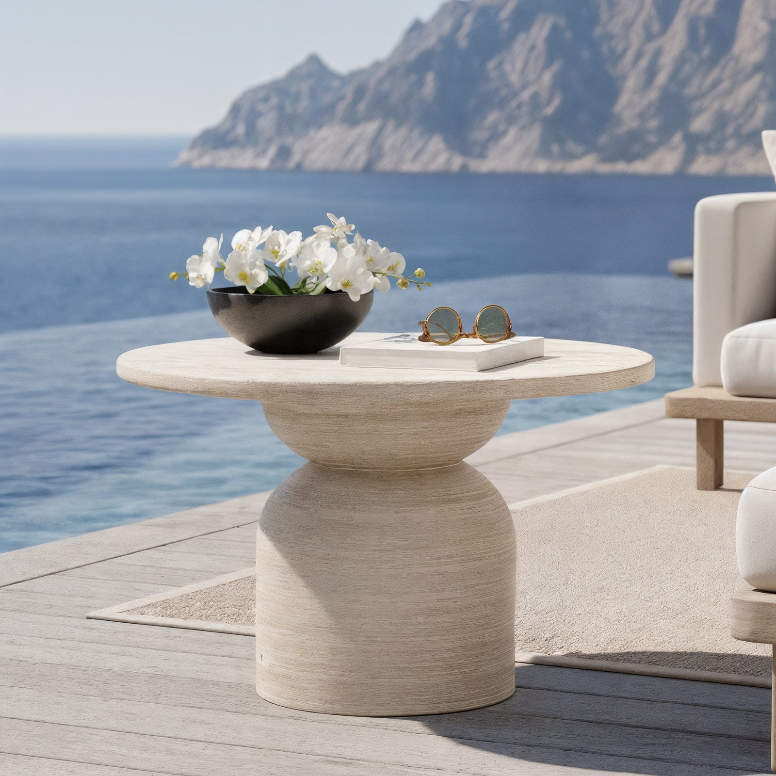 The image showcases a serene outdoor setting with a Krates 120cm Round Textured Outdoor Concrete Table in Beige by an infinity pool, complemented by a sculptural side table and coastal view.