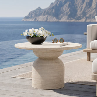 The image showcases a serene outdoor setting with a Krates 120cm Round Textured Outdoor Concrete Table in Beige by an infinity pool, complemented by a sculptural side table and coastal view.
