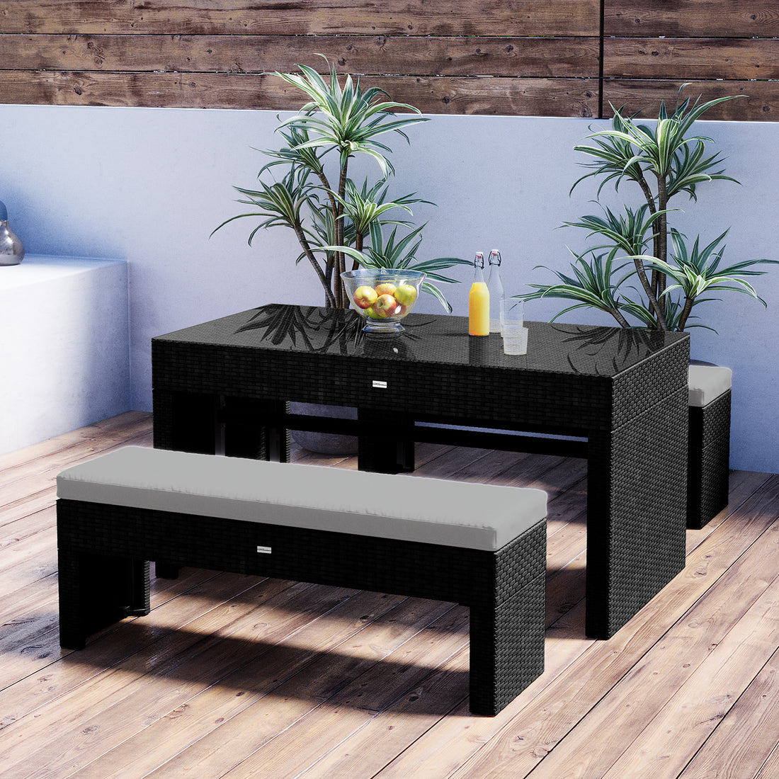 Kummi 4 Seater Outdoor Dining Bench Set features a black wicker table with glass top, apples, juice bottles, and glasses, paired with cushioned benches, and accented by potted tropical plants.