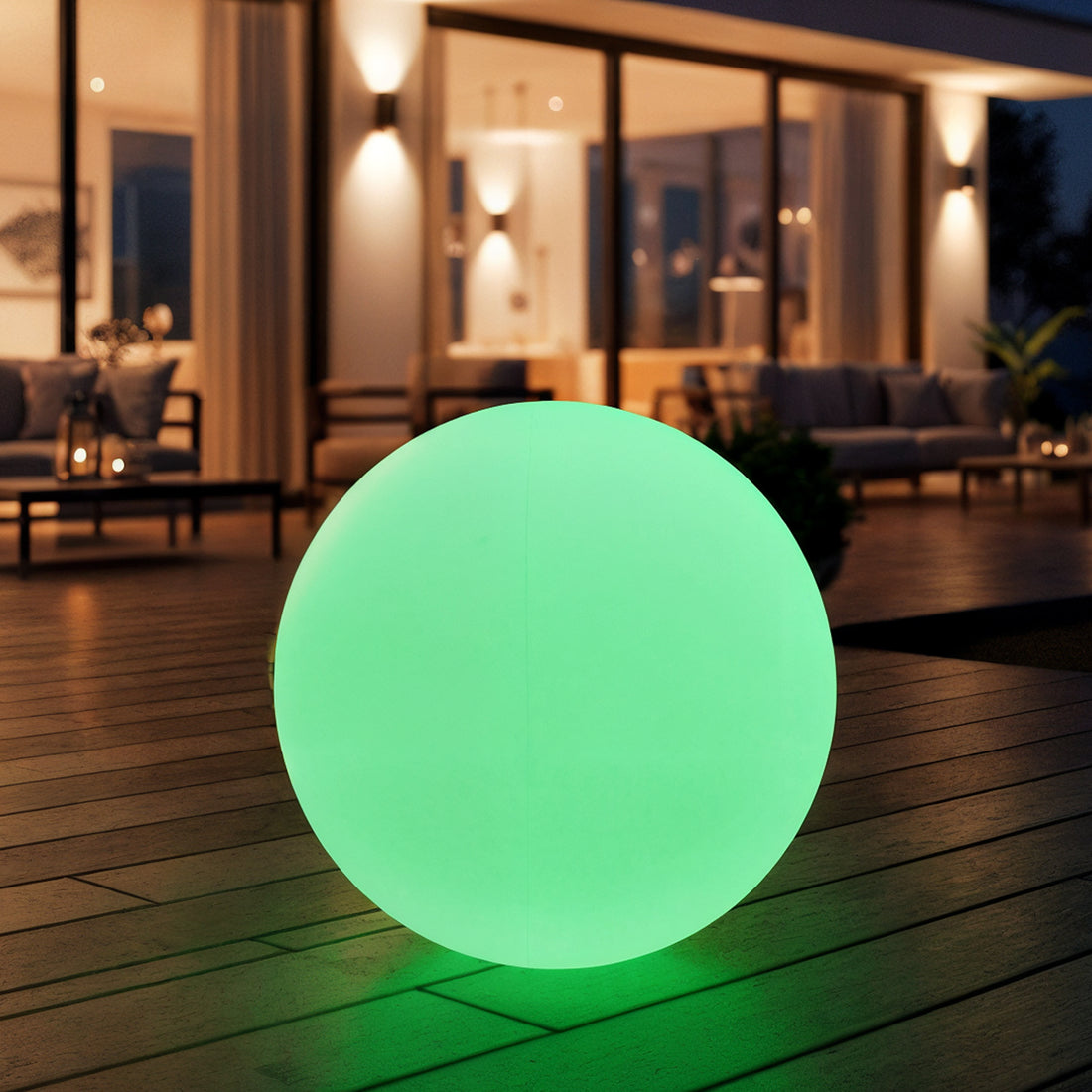 Sphero 20cm Floating Waterproof Outdoor LED Ball Lights on a wooden deck, glowing green against a modern house backdrop with floor-to-ceiling windows and a softly lit seating area at dusk.
