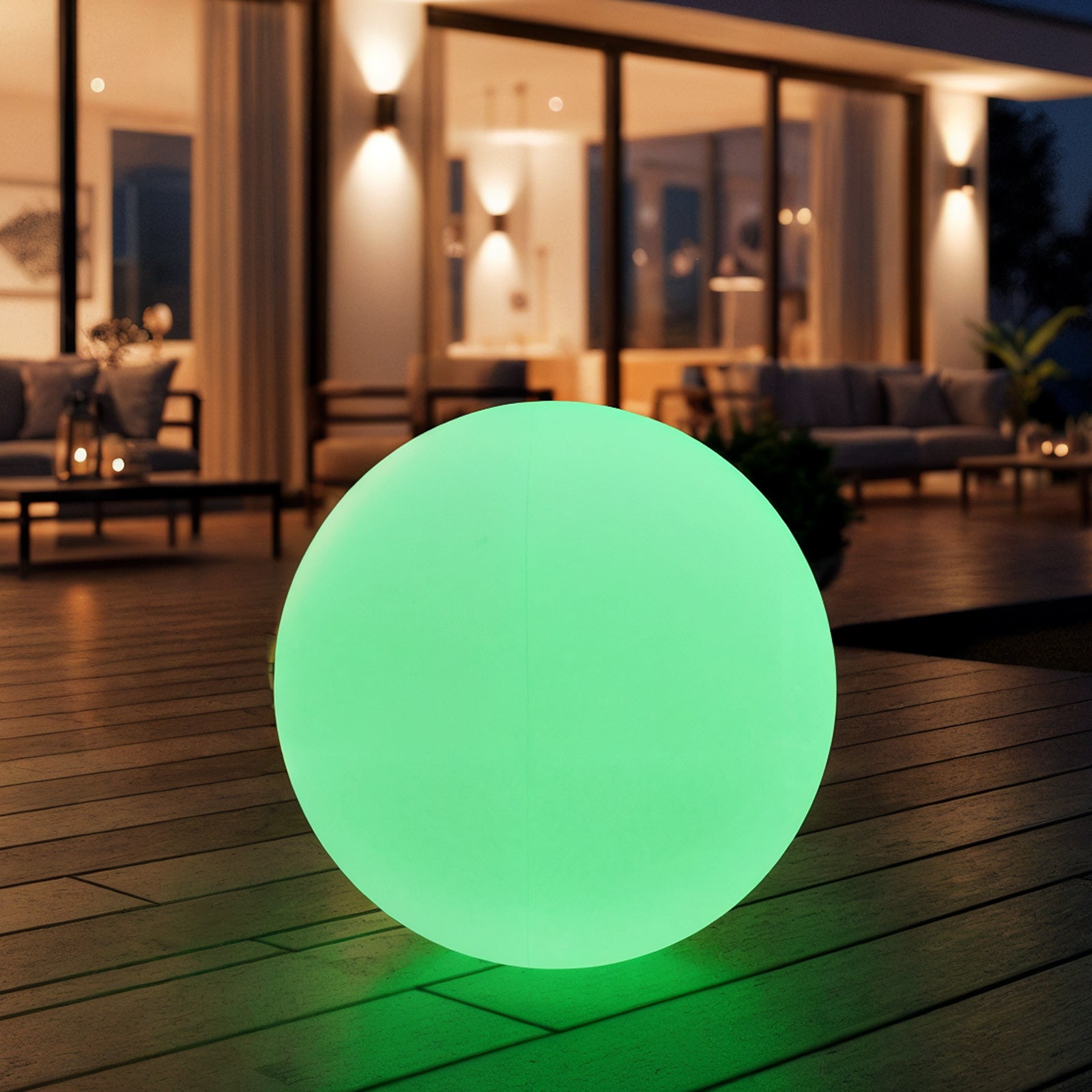 Sphero 20cm Floating Waterproof Outdoor LED Ball Lights on a wooden deck, glowing green against a modern house backdrop with floor-to-ceiling windows and a softly lit seating area at dusk.
