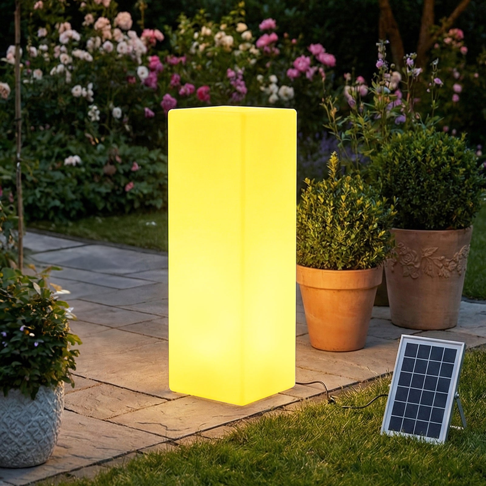 80cm Outdoor Solar LED Column Light with Remote Control on stone paving, glowing yellow at dusk, connected to a small solar panel on grass, surrounded by terracotta pots and flowering bushes.