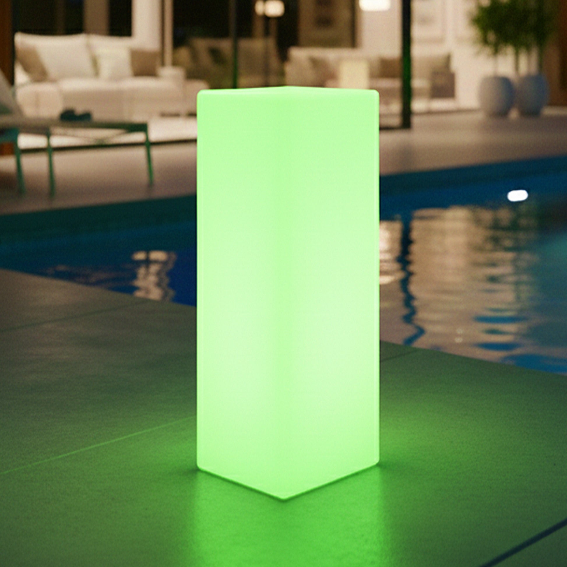 The 16 Colour‑Changing Outdoor Waterproof LED Column Light, glowing green, stands on a tiled floor by a pool, enhancing the modern indoor/outdoor lounge with ambient lighting.
