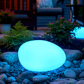 The 22cm Waterproof Solar Charging Outdoor LED Rock Light glows blue, resembling a smooth stone, set on pebbles in a garden, surrounded by low plants and patio furniture.