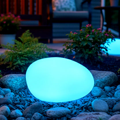 The 22cm Waterproof Solar Charging Outdoor LED Rock Light glows blue, resembling a smooth stone, set on pebbles in a garden, surrounded by low plants and patio furniture.
