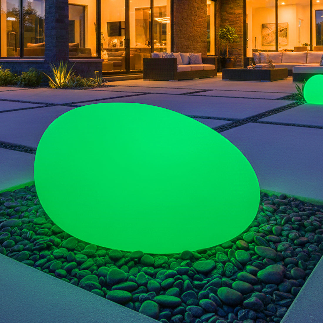 40cm Waterproof Solar Charging Outdoor LED Rock Light nestled in river stones, glowing in a modern patio scene at night with a contemporary home backdrop, enhancing outdoor ambience effortlessly.