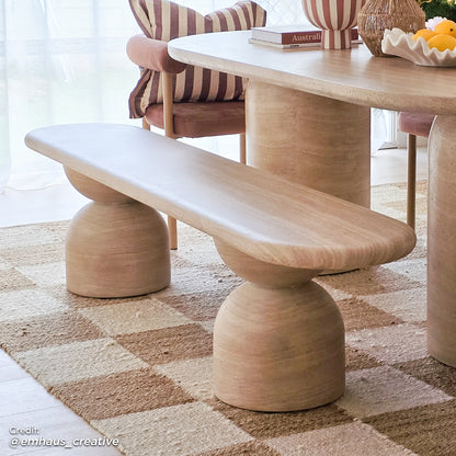 Lumen 200cm Oval Outdoor Concrete Bench in Beige features a minimalist design with a curved seat on half-sphere pedestal legs, set on a checkerboard-patterned rug beside a rounded table.