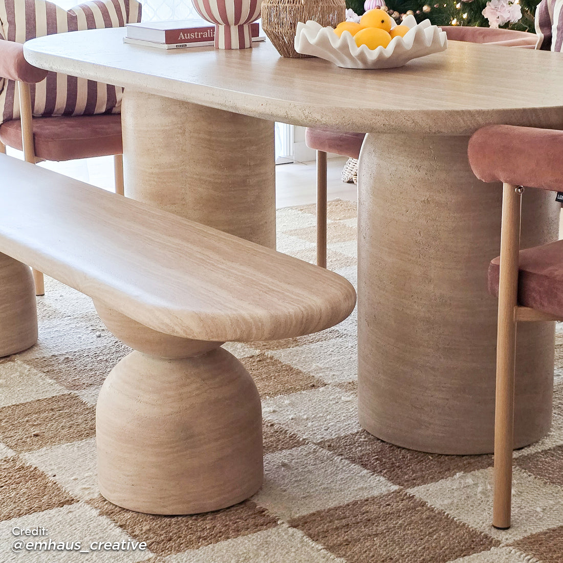 Lumen 220cm Outdoor Concrete Dining Table Set in Beige features an oval table with a sculptural base, matching bench, and rose-pink velvet chairs, set on a checker-patterned rug with decorative tabletop accents.