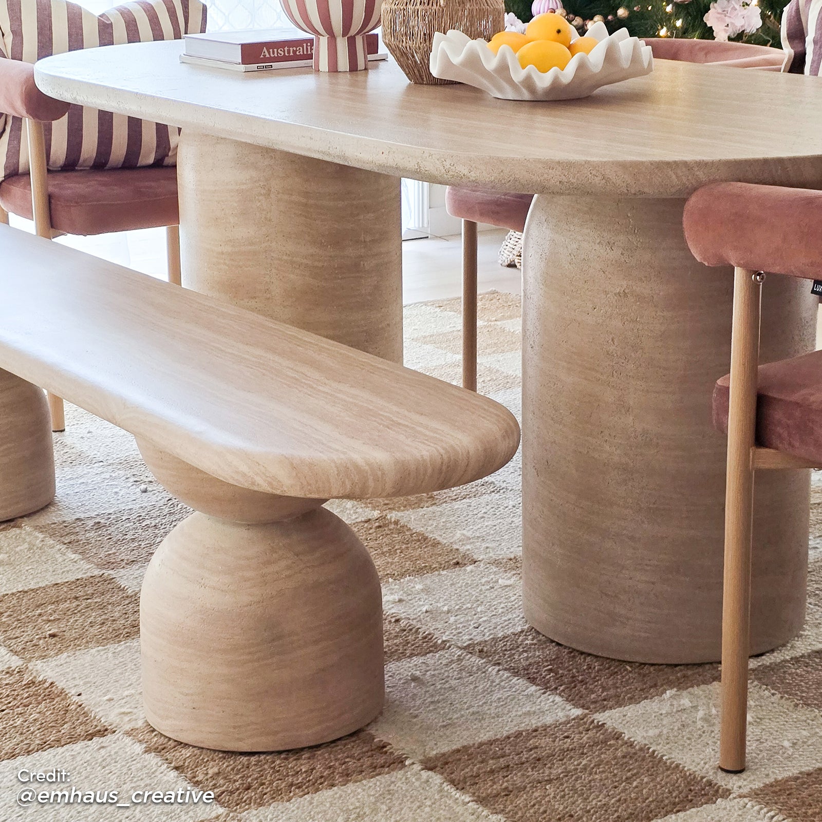 Lumen 220cm Outdoor Concrete Dining Table Set in Beige features an oval table with a sculptural base, matching bench, and rose-pink velvet chairs, set on a checker-patterned rug with decorative tabletop accents.