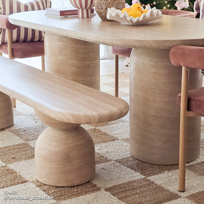 Lumen 220cm Outdoor Concrete Dining Table Set in Beige features an oval table with a sculptural base, matching bench, and rose-pink velvet chairs, set on a checker-patterned rug with decorative tabletop accents.