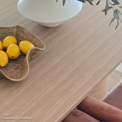 Lumen 220cm Oval Outdoor Concrete Dining Table in Beige with a sleek, oval top and cone-shaped base, surrounded by wicker bowl of lemons and white vase with green branches on a light wood tabletop.