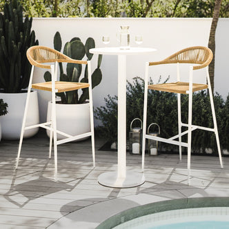 Luzon & Dinagat Wicker Outdoor Bar Table Set in Natural, featuring a white bistro table with a glass pitcher and two bar stools with woven natural-fiber seats, set by a poolside.