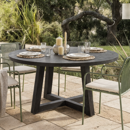 Magnus Hardwood and Concrete Outdoor Dining Table on a patio with a round concrete top, acacia wood X-base, set with rattan placemats and surrounded by green metal chairs with cushions.