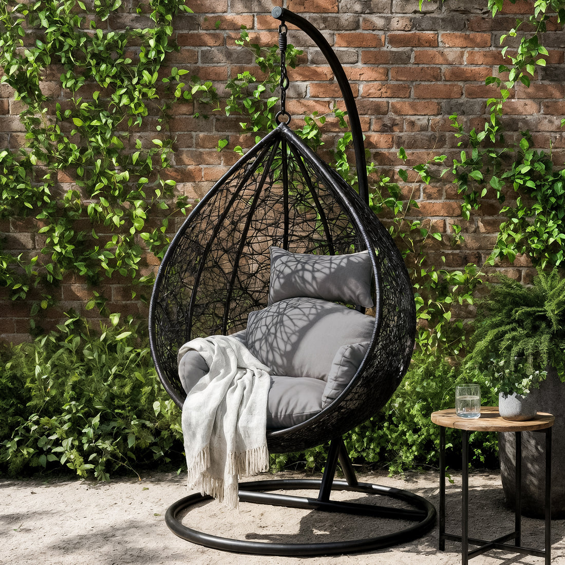 Marian Large Hanging Outdoor Egg Chair Black in a garden setup, featuring a sleek black-metal frame, woven shell with thick cushions, a draped throw blanket, and a nearby wooden side table with plant.