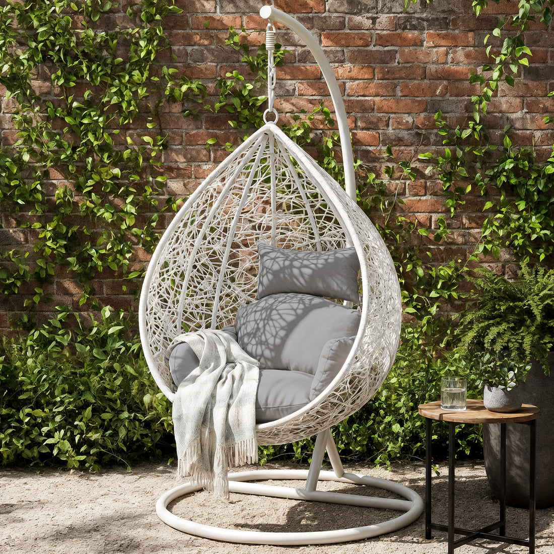 Marian Large Hanging Outdoor Egg Chair White, featuring a cozy cocoon design with plush grey cushions, suspended on a stand. Accompanied by a side table with a plant, set against a verdant garden backdrop.