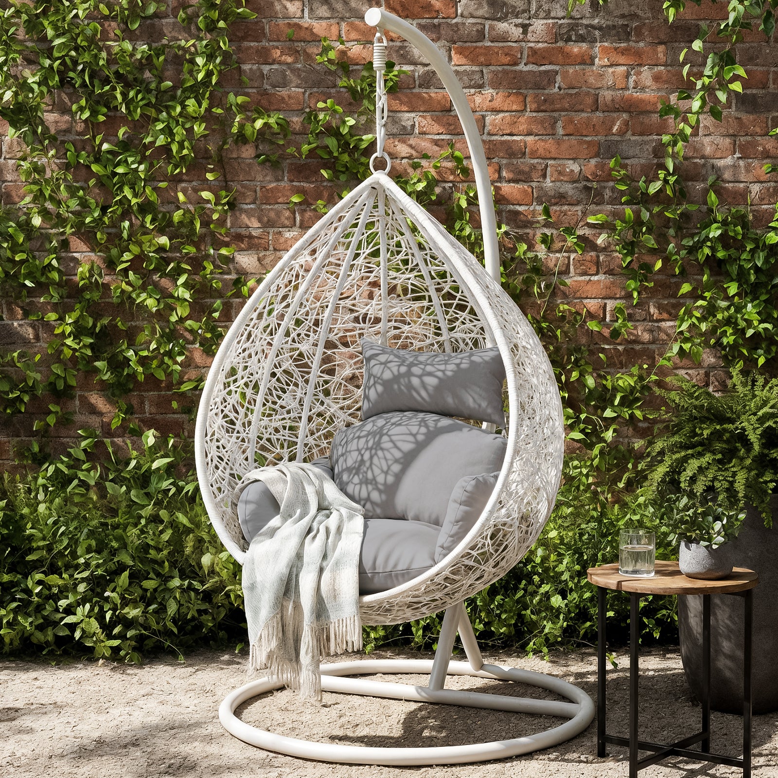 Marian Large Hanging Outdoor Egg Chair White, featuring a cozy cocoon design with plush grey cushions, suspended on a stand. Accompanied by a side table with a plant, set against a verdant garden backdrop.