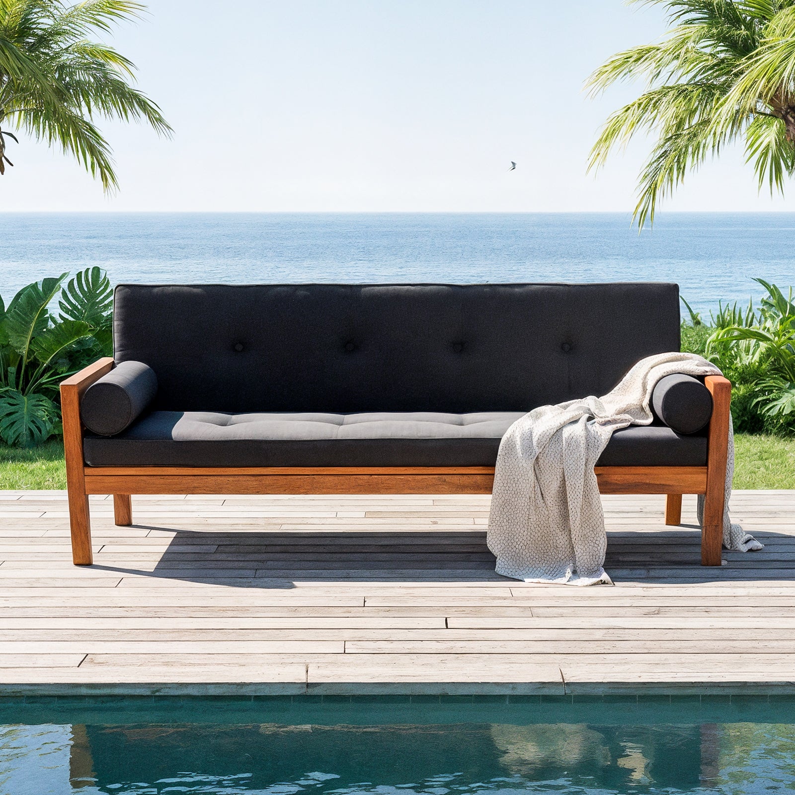 Luxo Meves Hardwood Outdoor Daybed Sofa & Cushion in Black, featuring a wooden frame with dark gray cushions and bolster pillows, set on a deck beside a pool with tropical ocean view.