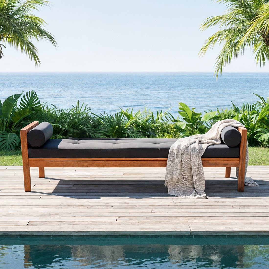 Luxo Meves Hardwood Outdoor Daybed Sofa & Cushion in Black, featuring a teak-like frame, dark gray cushions with bolsters, and a draped throw, set on a poolside deck amidst tropical greenery.