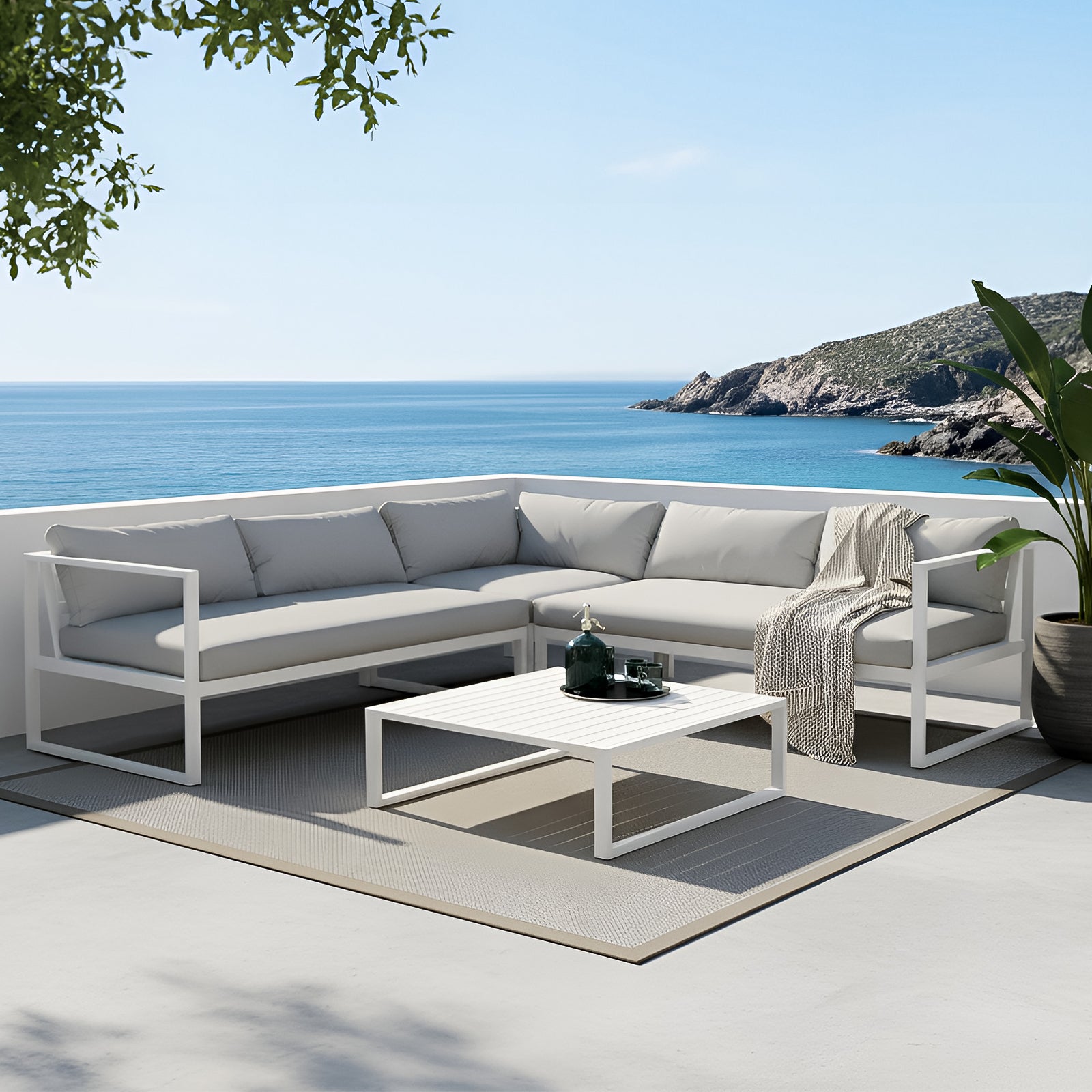 Moana 6-Seater Corner Outdoor Sofa Set in Light Grey on a sunlit seaside terrace, featuring a white-framed L-shaped sectional with pale gray cushions and a matching coffee table, complemented by dappled shadows and sea views.
