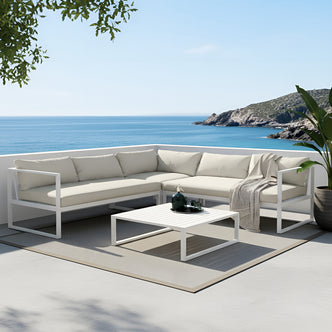 Moana 6 Seater Corner Outdoor Sofa Set in Sandy Beige features an L-shaped design with a white metal frame, pale-beige cushions, a low square coffee table, and a decorative vase on a coastal terrace.