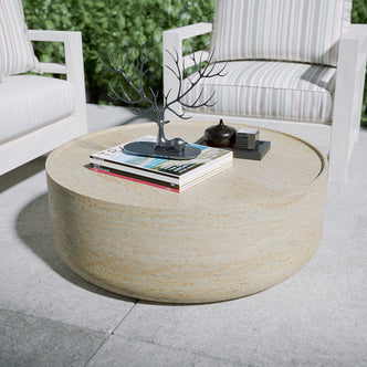 Myrtle 80cm Concrete Indoor/Outdoor Coffee Table in Beige, carved from concrete, holds a magazine stack, decorative deer-shaped stand, small lidded vessel, and tray on a tiled patio with striped-cushioned chairs.