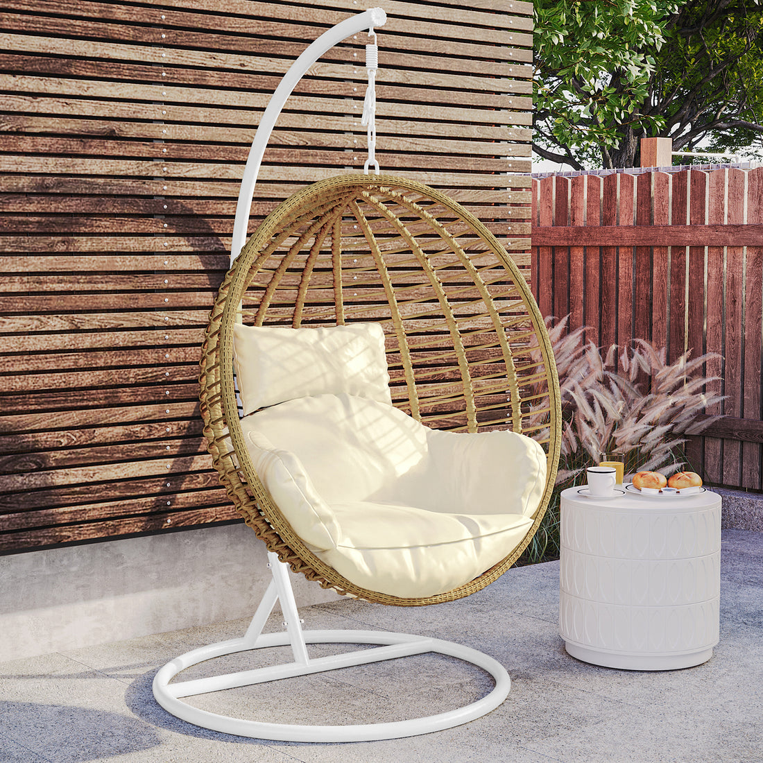 Napili Outdoor Rattan Egg Chair with Cushion in Cream White, featuring a suspended woven rattan design, thick cushion, and matching back pillow, situated beside a small white side table with refreshments.