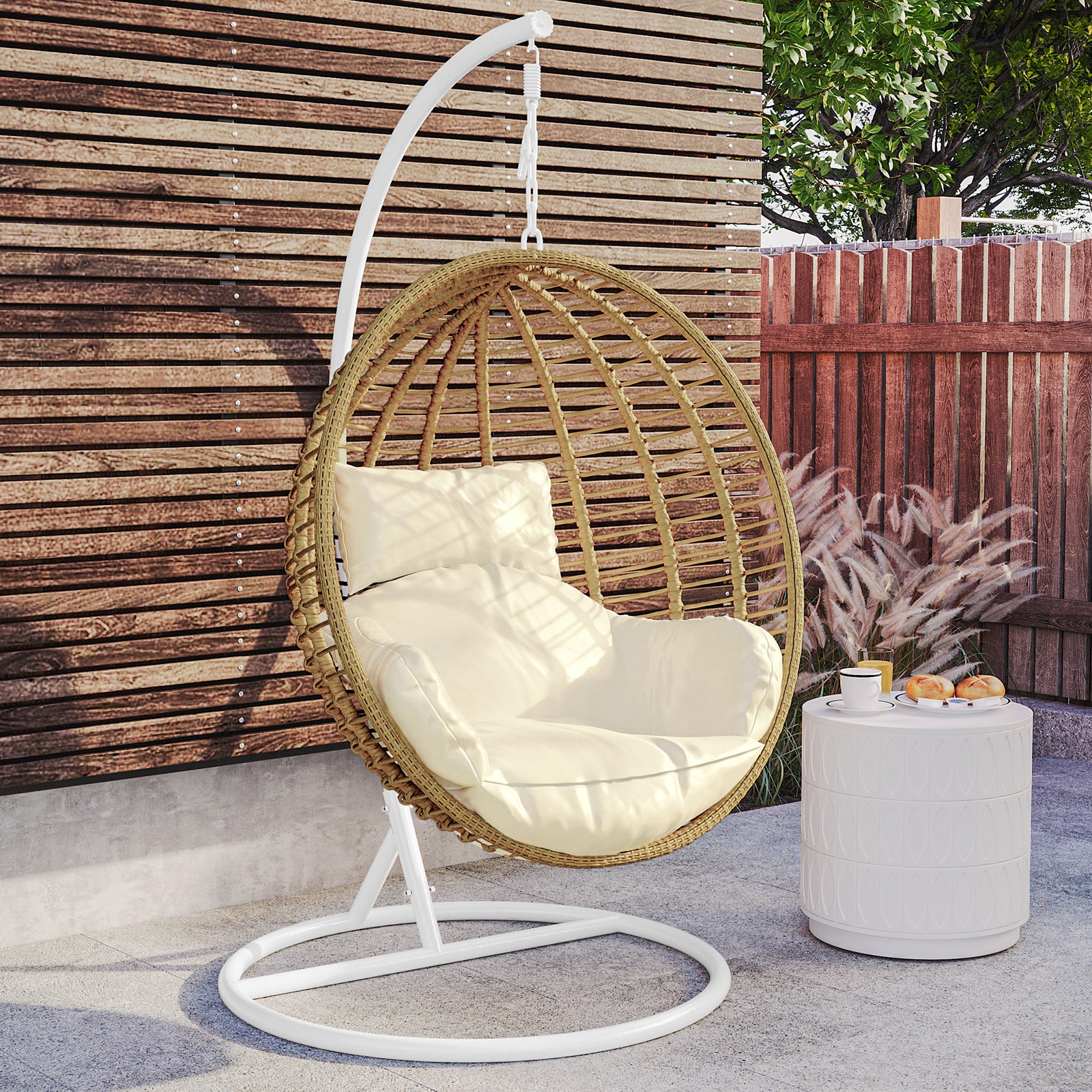 Napili Outdoor Rattan Egg Chair with Cushion in Cream White, featuring a suspended woven rattan design, thick cushion, and matching back pillow, situated beside a small white side table with refreshments.