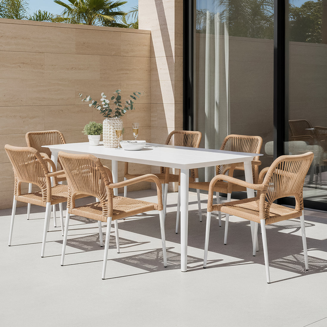 Navarre 6 Seater Aluminium Outdoor Dining Set in Natural features a white table with six wicker-style chairs, complemented by a ceramic vase, potted plant, glasses, and a serving bowl on a patio.