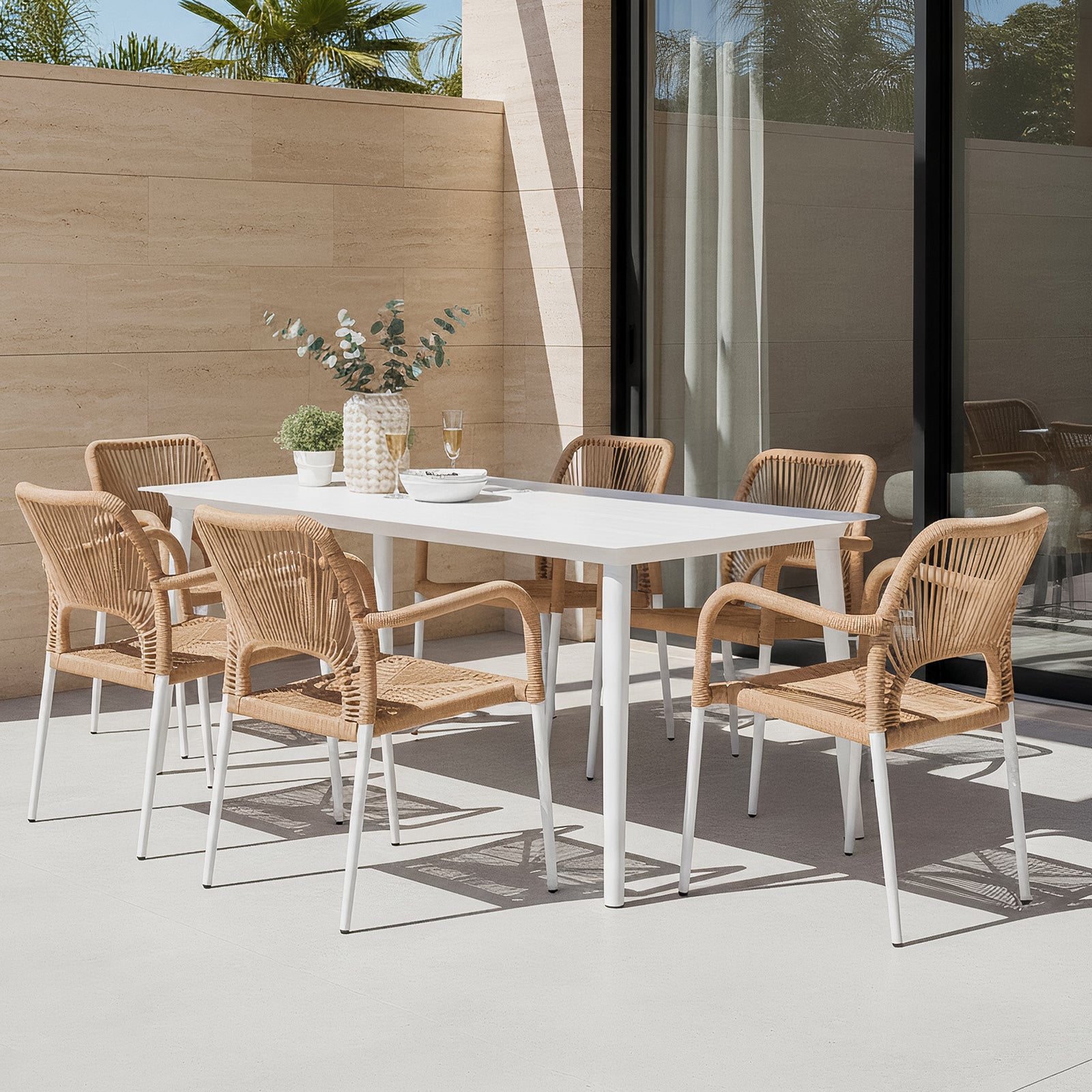 Navarre 6 Seater Aluminium Outdoor Dining Set in Natural features a white table with six wicker-style chairs, complemented by a ceramic vase, potted plant, glasses, and a serving bowl on a patio.