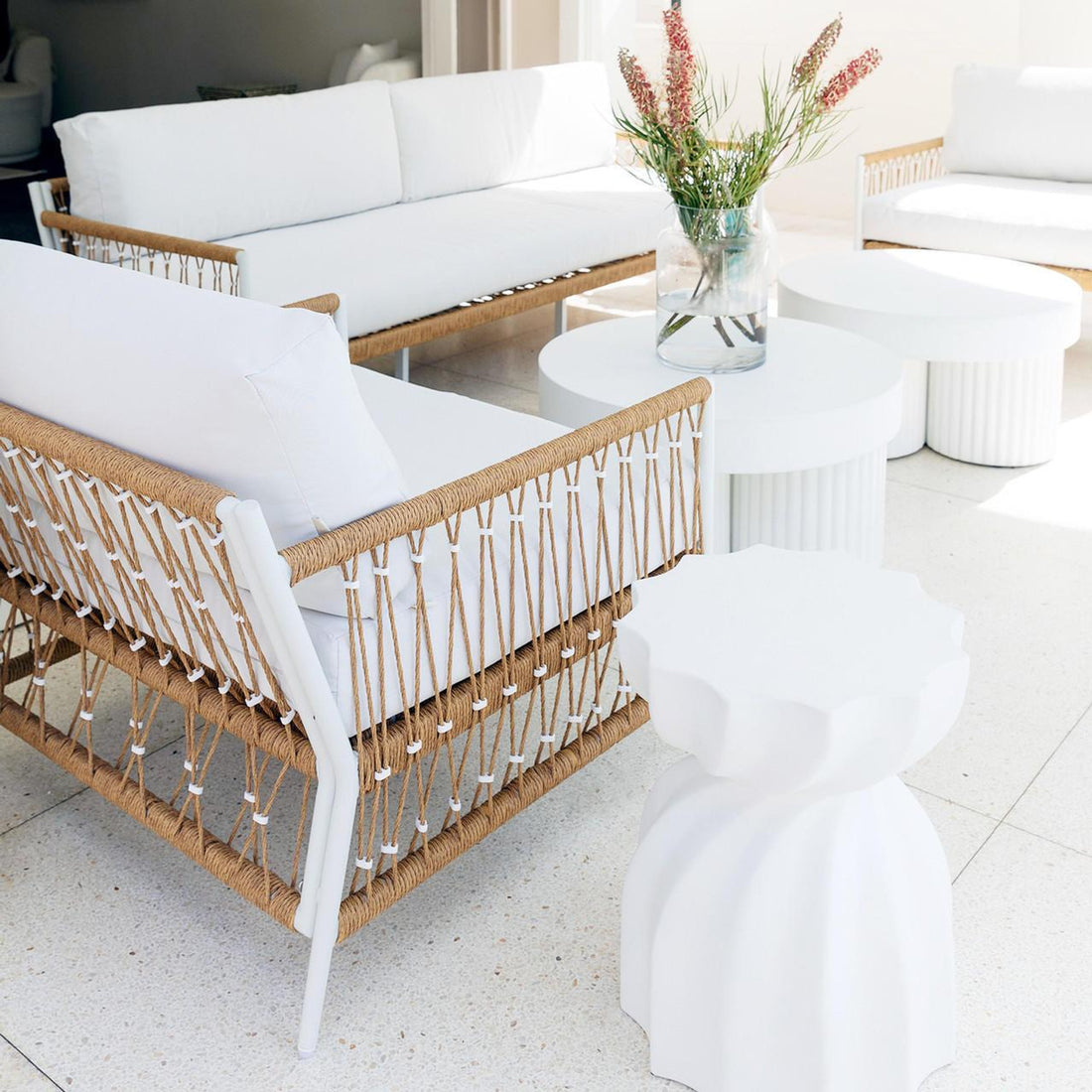 Nuevo 4 Seater Wicker Outdoor Lounge Set in Light Beige, featuring white-upholstered sofas with white metal frames and natural rope weaving, encircling small round tables with a vase of vibrant red and green blooms.