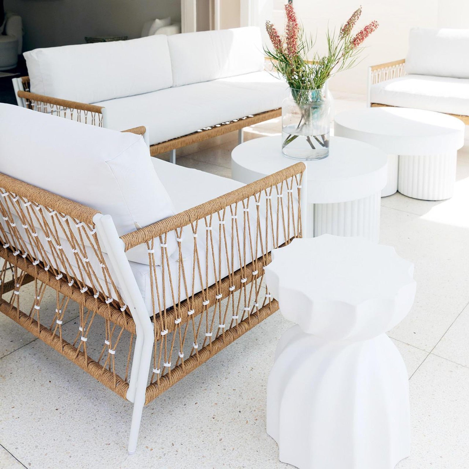 Nuevo 4 Seater Wicker Outdoor Lounge Set in Light Beige, featuring white-upholstered sofas with white metal frames and natural rope weaving, encircling small round tables with a vase of vibrant red and green blooms.