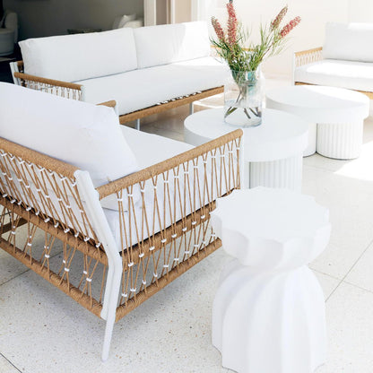 Nuevo 4 Seater Wicker Outdoor Lounge Set in Light Beige, featuring white-upholstered sofas with white metal frames and natural rope weaving, encircling small round tables with a vase of vibrant red and green blooms.