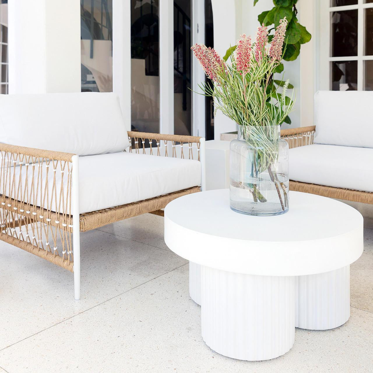 Nuevo 4 Seater Wicker Outdoor Lounge Set in Light Beige featuring two lounge chairs with woven-rope frames and white cushions, complemented by a sculptural white coffee table with pink flowers in a vase.