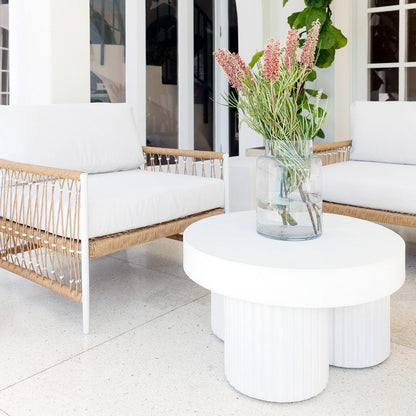 Nuevo 4 Seater Wicker Outdoor Lounge Set in Light Beige featuring two lounge chairs with woven-rope frames and white cushions, complemented by a sculptural white coffee table with pink flowers in a vase.