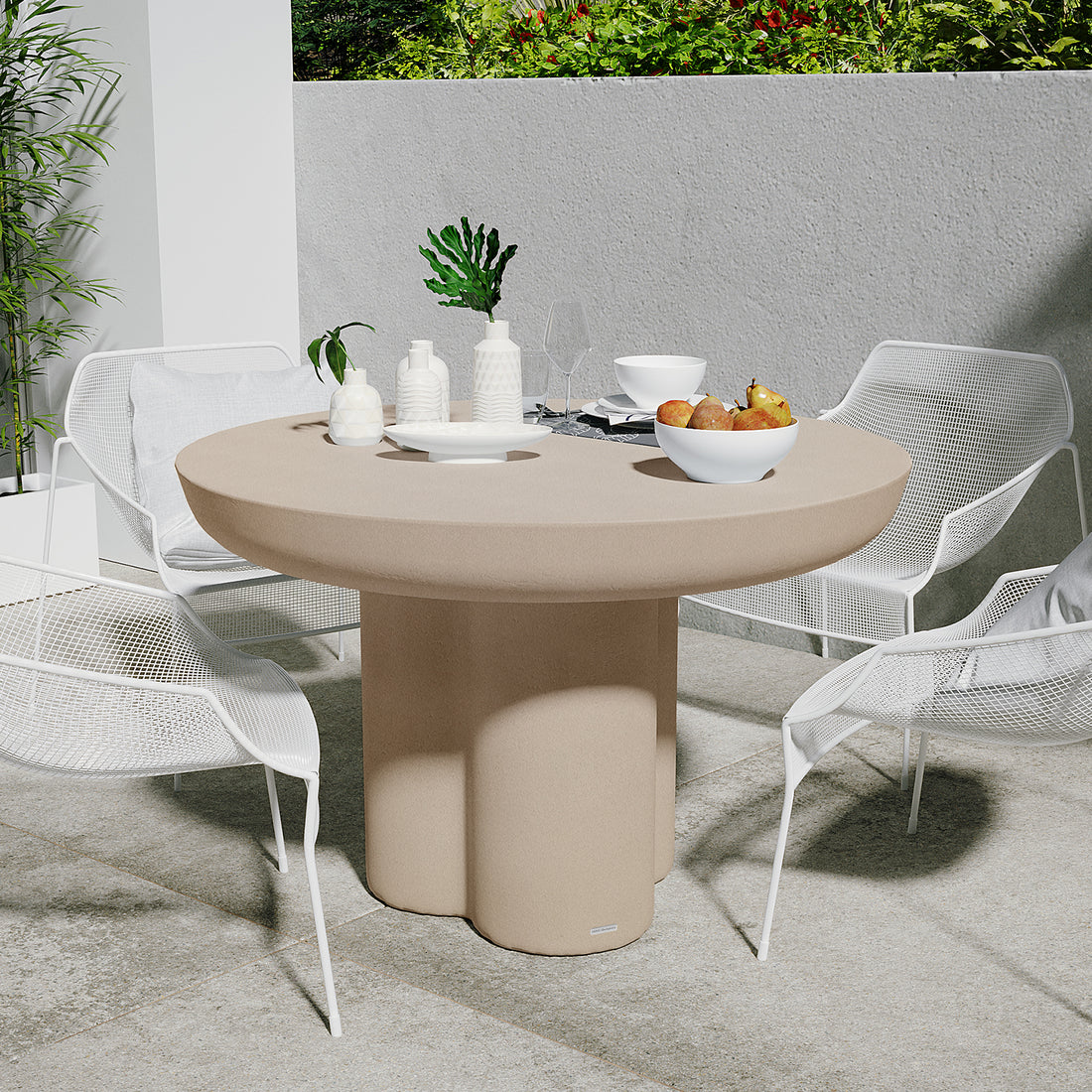 Oberon 120cm Round Outdoor Concrete Dining Table Ivory with a white bowl of fruit, ceramic vases, and dining essentials. Surrounded by four white mesh chairs on a stone-tiled patio with greenery.