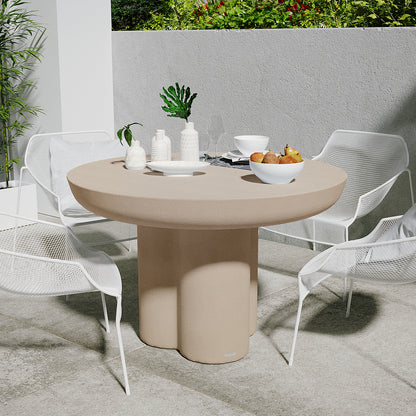 Oberon 120cm Round Outdoor Concrete Dining Table Ivory with a white bowl of fruit, ceramic vases, and dining essentials. Surrounded by four white mesh chairs on a stone-tiled patio with greenery.