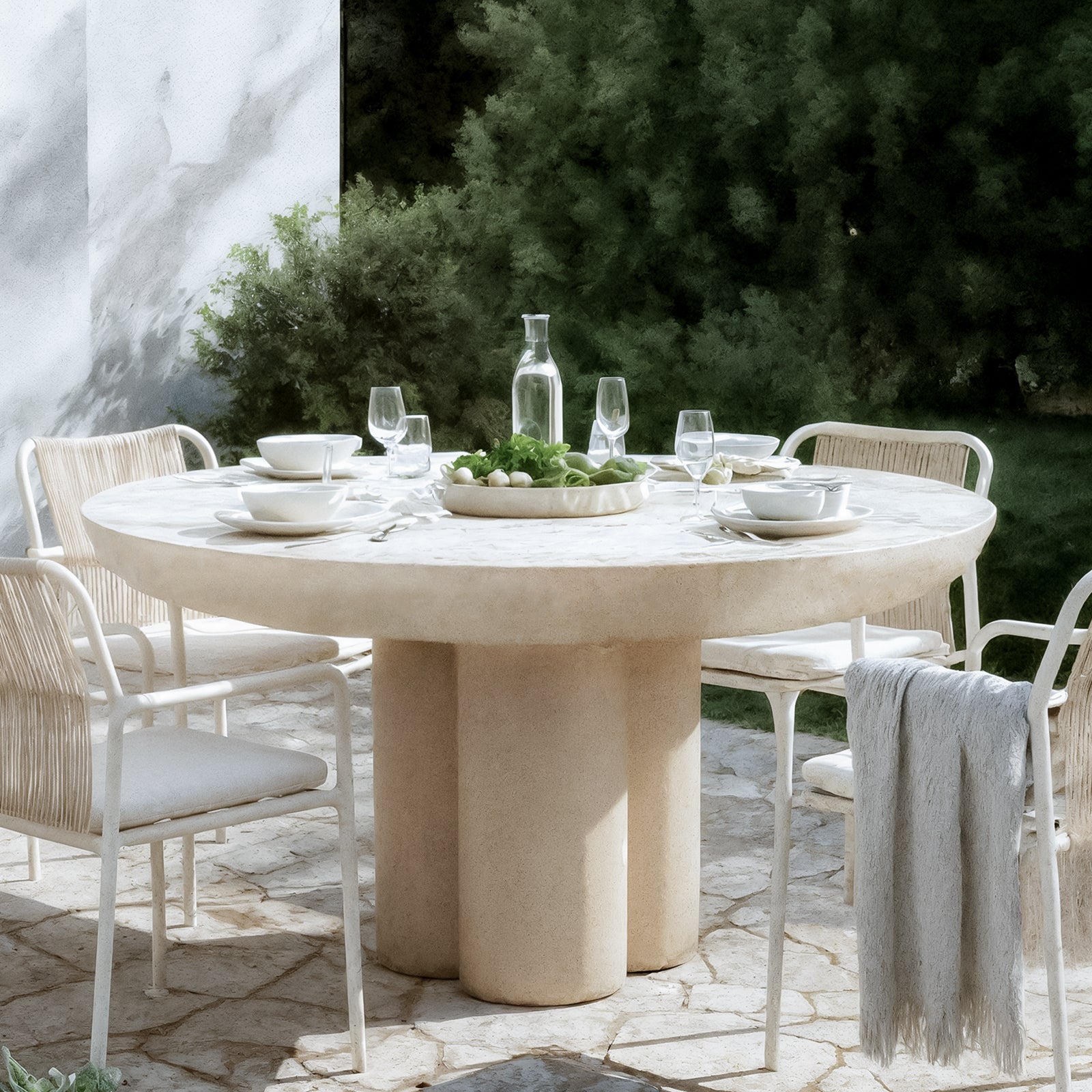 Oberon 120cm Round Outdoor Concrete Dining Table in Ivory features a sculptural three-pillar base, surrounded by white metal-frame chairs, set with elegant dining ware amidst lush greenery.