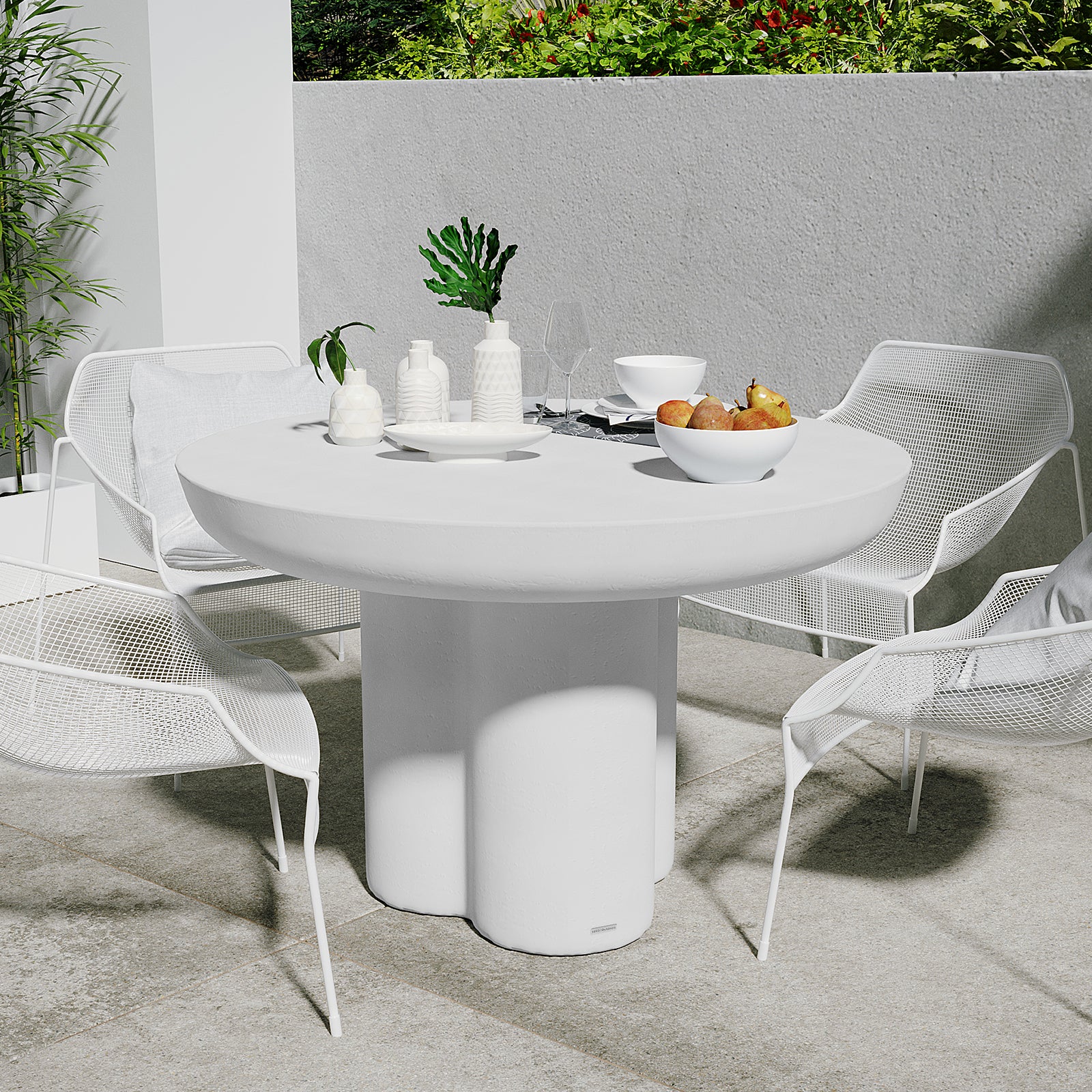 Oberon 120cm Round Outdoor Concrete Dining Table White, with a sculptural pedestal base, surrounded by four metal-mesh chairs, adorned with white ceramic pieces, greenery, and a bowl of pears.