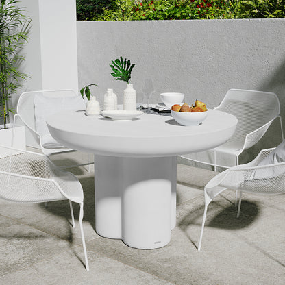 Oberon 120cm Round Outdoor Concrete Dining Table White, with a sculptural pedestal base, surrounded by four metal-mesh chairs, adorned with white ceramic pieces, greenery, and a bowl of pears.
